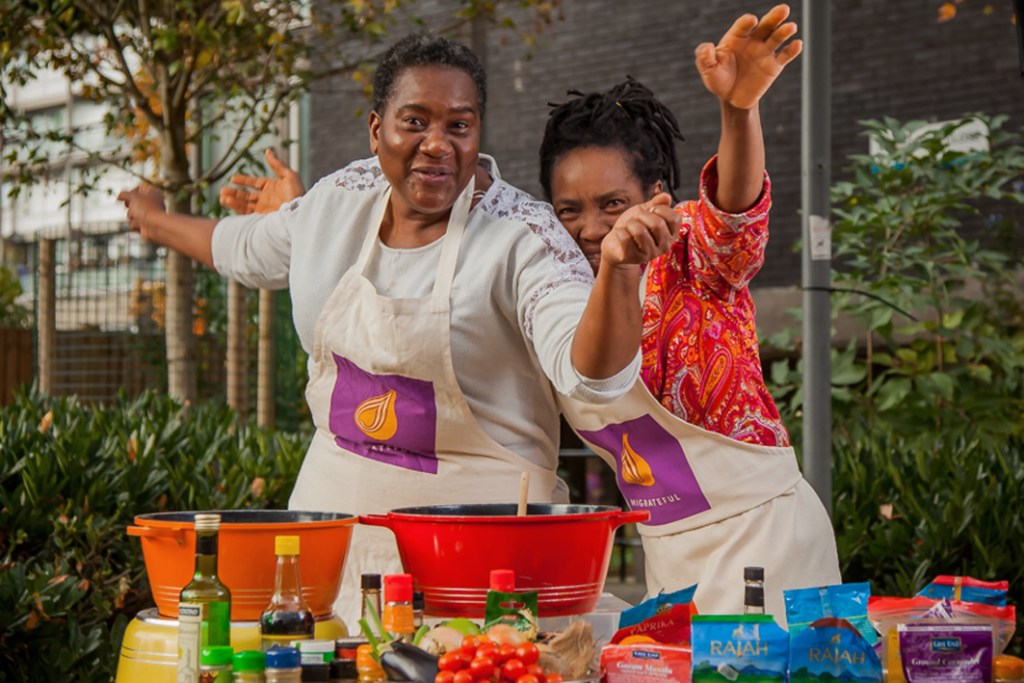 Two black women wearing a white apron with the purple and orange Migrateful logo, standing in front of a table with pots and cooking ingredients