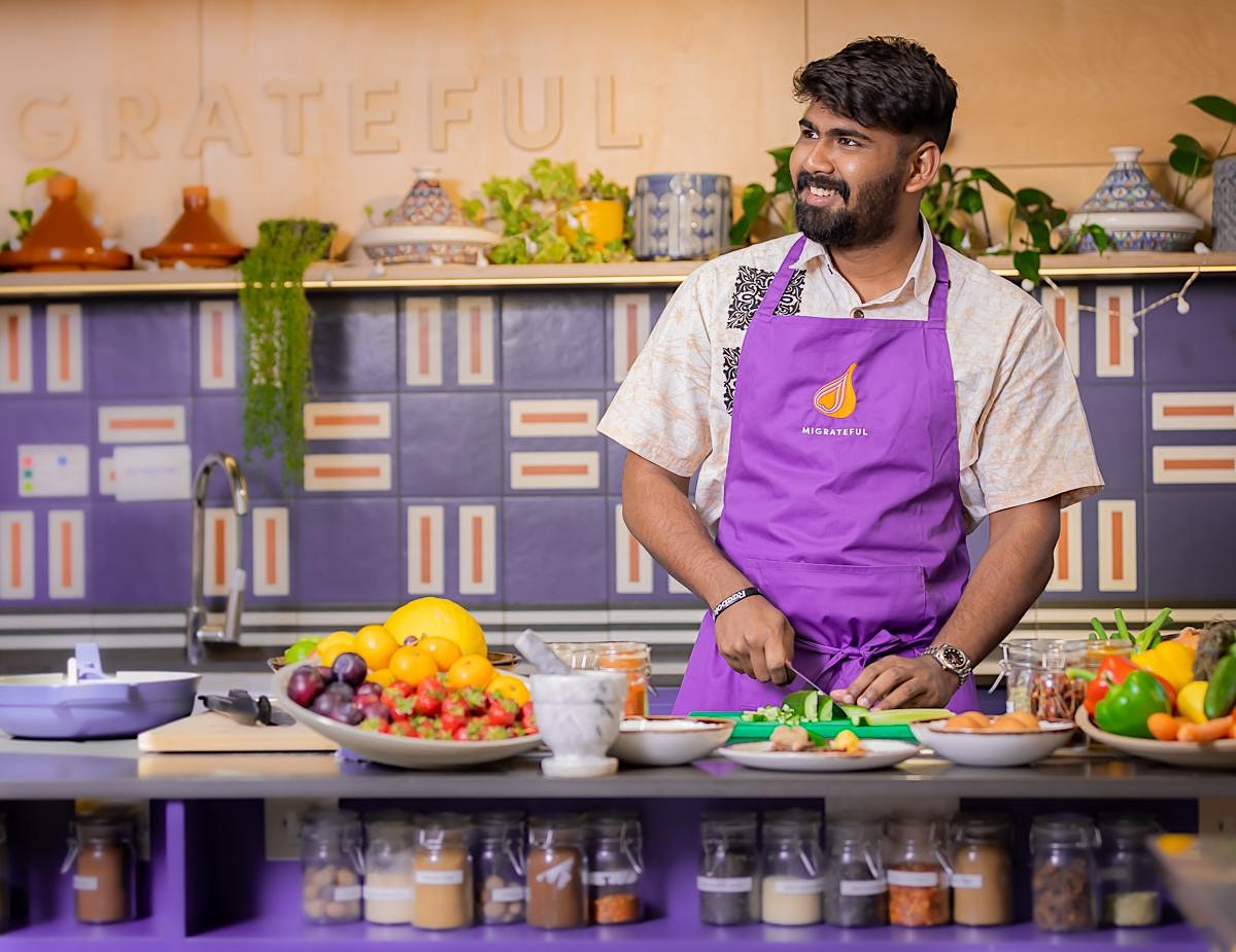 A man in an apron is preparing food in a purple kitchen as part of the Migrateful London Cookery Class.