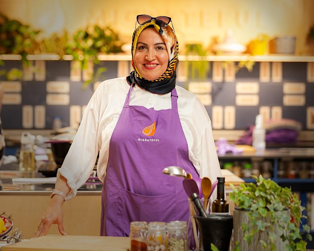 A woman in a purple apron standing in a kitchen, passionately engaging in a London Cookery Class offered by the organization Migrateful.
