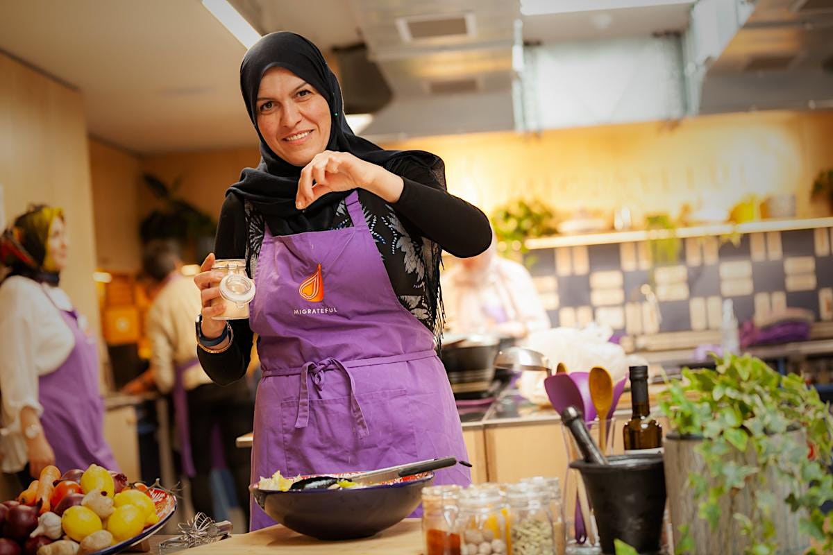 A Migrateful London Cookery Class instructor, dressed in a vibrant purple apron, skillfully prepares food in a bustling kitchen.