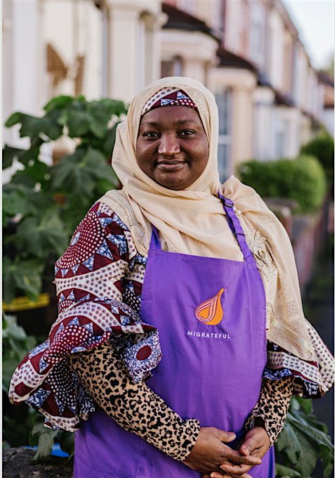 A woman in a purple apron standing in front of a house, representing a London Cookery Class.