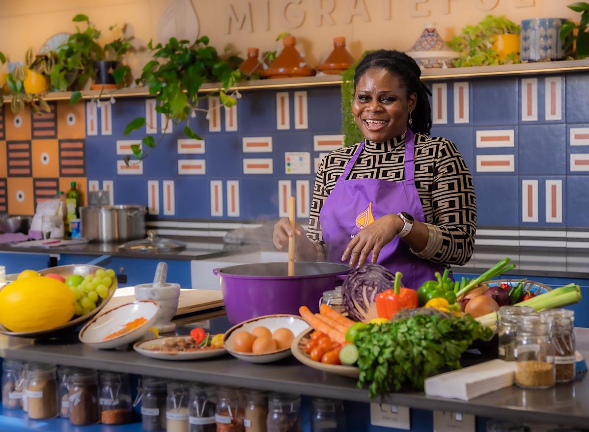 A Migrateful London Cookery Class instructor, wearing an apron, is standing in front of a kitchen full of vegetables.