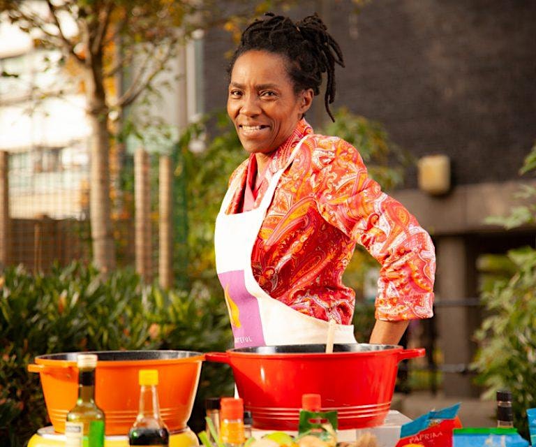 A woman with dreadlocks smiling in front of colorful pots while participating in a Migrateful London Cookery Class.
