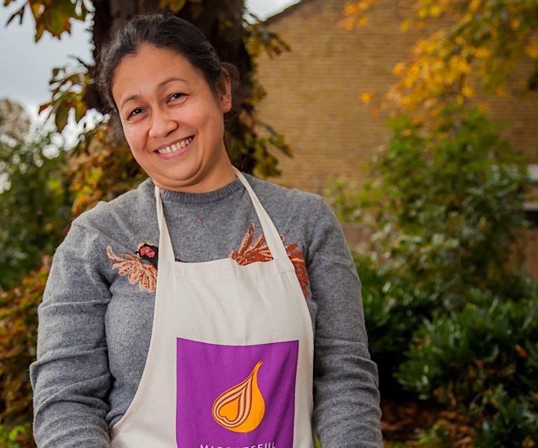 A woman with a purple apron smiling in front of a tree, attending a London Cookery Class organized by Migrateful.