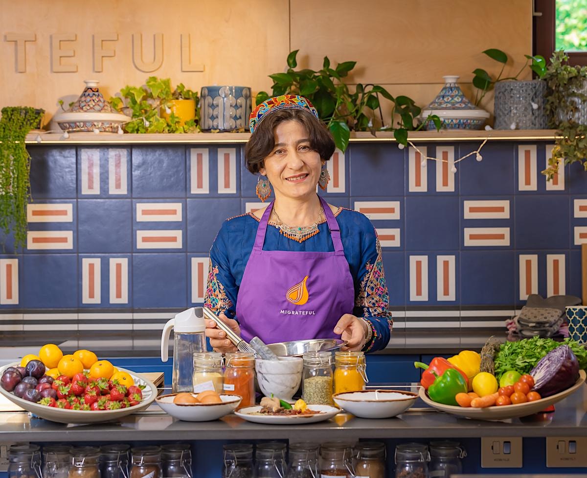A Migrateful London Cookery Class instructor, wearing an apron, stands proudly in front of a kitchen full of delicious food.