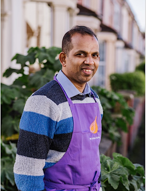 A man in a purple apron standing in front of a house, representing the London Cookery Class offered by Migrateful.