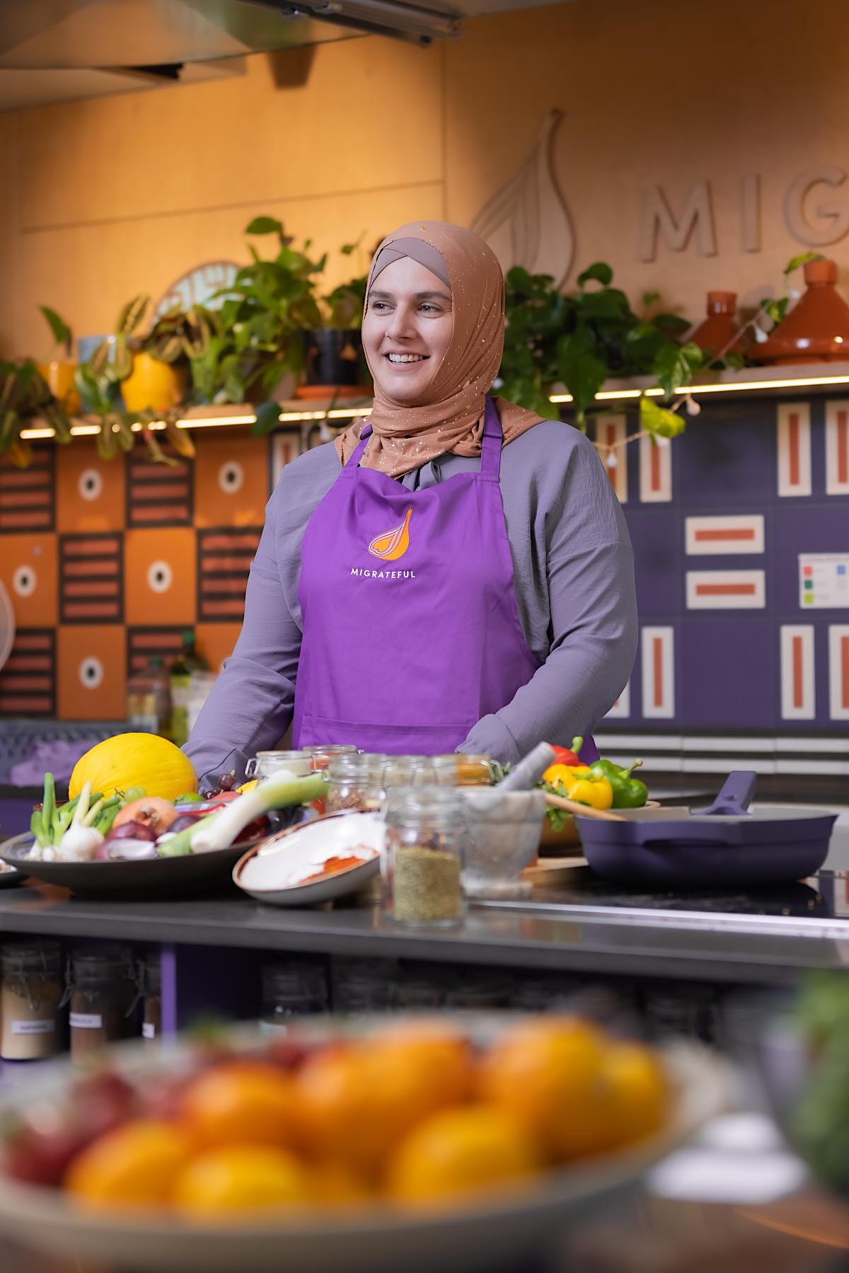 A woman in a purple apron standing in front of a kitchen at a London Cookery Class.